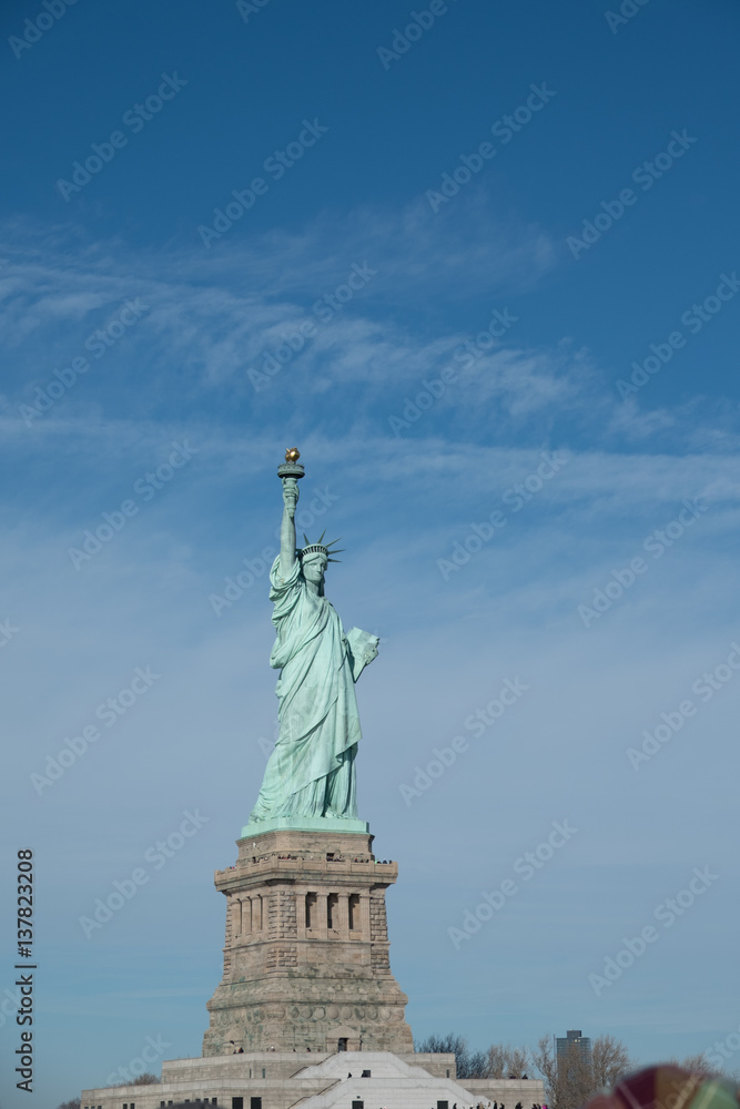 Obraz premium Statue of Liberty on pedestal on Liberty Island, view from passing ferry. Blue skies, wispy clouds on a sunny day in New York. Lady Liberty overlooks New York City Harbor, top New York sights.