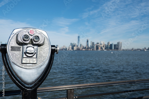 Tower viewer binoculars on a swivel stand next to a railing on the edge of Liberty Island, New York City.  View of Lower Manhattan and New York Harbor from the Statue of Liberty. Bright, sunny day.