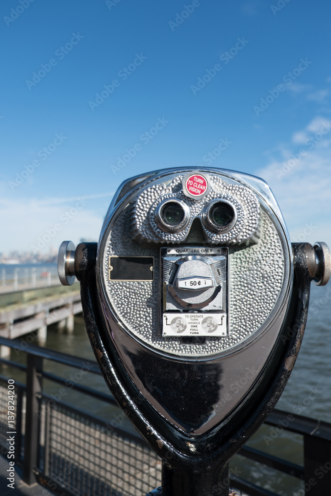 Tower viewer binoculars on Liberty Island looking towards Manhattan, New York. Pier and Manhattan skyline in the distance. Shallow depth of field, focus on telescoping tower viewer in the foreground. Tower viewer binoculars on Liberty Island looking towards Manhattan, New York. Pier and Manhattan skyline in the distance. Shallow depth of field, focus on telescoping tower viewer in the foreground.
