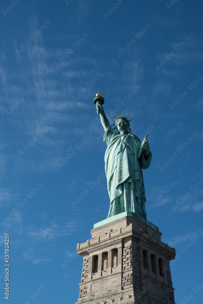 Fototapeta premium Statua Wolności na piedestale z błękitnym niebem i cienkimi chmurami, Liberty Island, Nowy Jork, USA w jasny słoneczny zimowy dzień. Żadnych ludzi. Lady Liberty spoglądająca w stronę portu w Nowym Jorku.