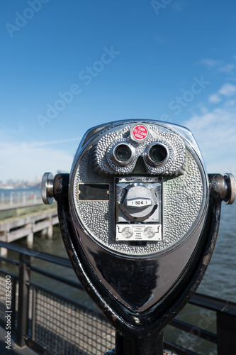 Tower viewer binoculars on Liberty Island looking towards Manhattan, New York. Pier and Manhattan skyline in the distance. Shallow depth of field, focus on telescoping tower viewer in the foreground.