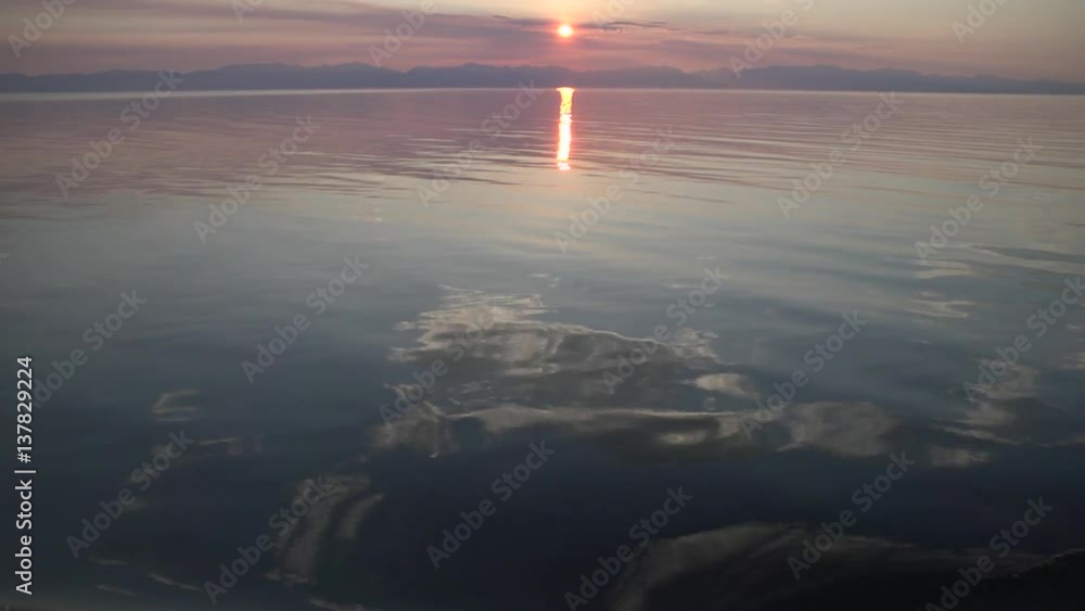 View from a moving boat on a summer sunset on the lake Baikal, Russia. The glare of the sun on the calm surface of the water. View from a moving boat on a summer sunset on the lake Baikal, Russia. The glare of the sun on the calm surface of the water.