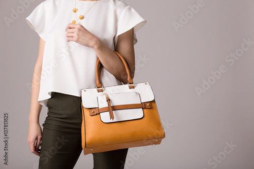 Stylish woman in modern clothes with bright orange white handbag in hands posing at studio isolated on gray background