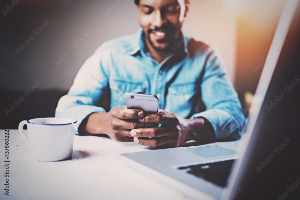 Happy African man using smartphone while sitting at the wooden table his modern home.Concept of young people working with mobile devices.Selective focus on male hands.Blurred background,flares. Happy African man using smartphone while sitting at the wooden table his modern home.Concept of young people working with mobile devices.Selective focus on male hands.Blurred background,flares.