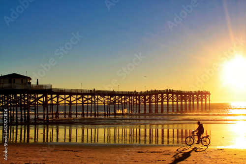 Bicyclist rides in the shadows and orange colors of a stunning San Diego sunset in California, with pier in background.