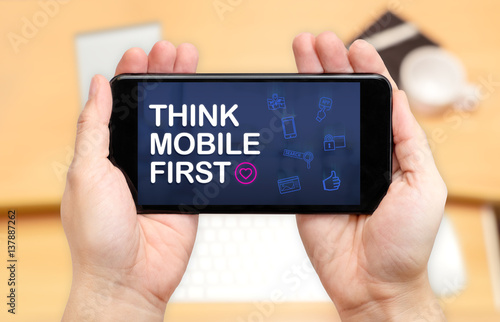 Watching two hand holding mobile phone with Think mobile first word and feature icon with blur desk office background,Digital marketing photo