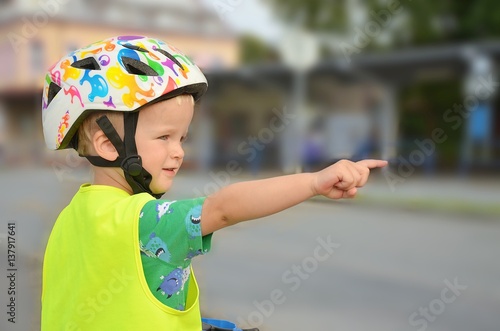 Little boy in the reflective vest pointing on the cars in the street. He is watching a traffic on the road