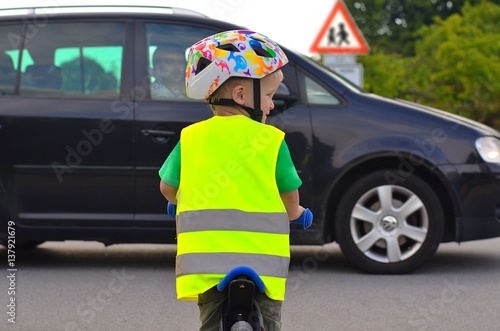 Little boy riding a bike and wearing reflective vest and helmet on the road. Driving car in front of him.