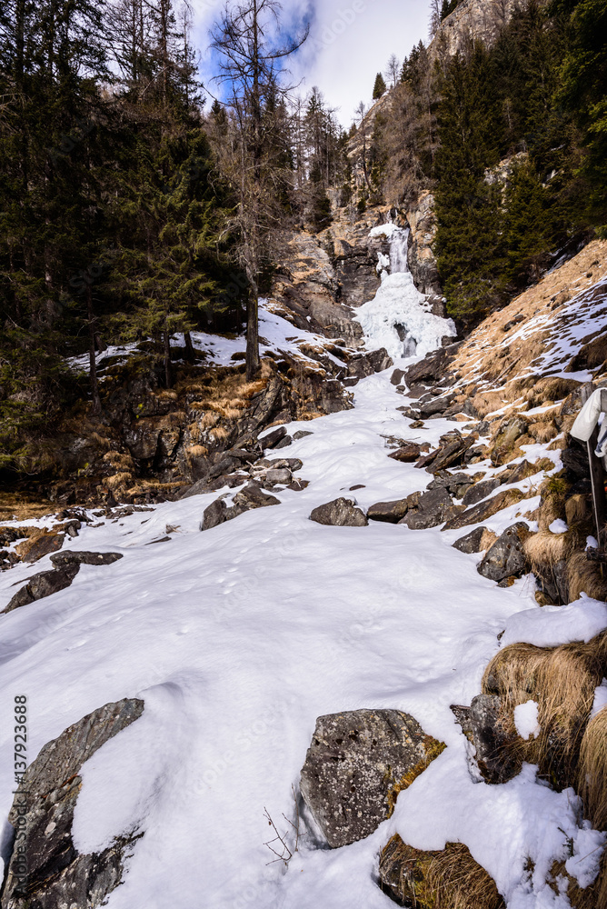 Saent waterfall, in Val di Rabbi, Trentino Alto Adige, covered by snow in a day with clear sky with few clouds. Trees are without snow. It a typical winter / spring mountain landscape/scenery Saent waterfall, in Val di Rabbi, Trentino Alto Adige, covered by snow in a day with clear sky with few clouds. Trees are without snow. It a typical winter / spring mountain landscape/scenery