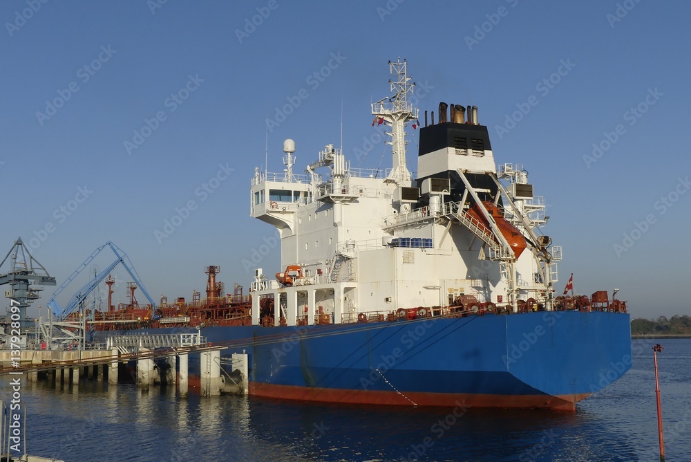 Products Tanker discharging at the Oil Terminal with blue hull and red main deck on a sunny day.