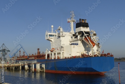 Products Tanker discharging at the Oil Terminal with blue hull and red main deck on a sunny day.