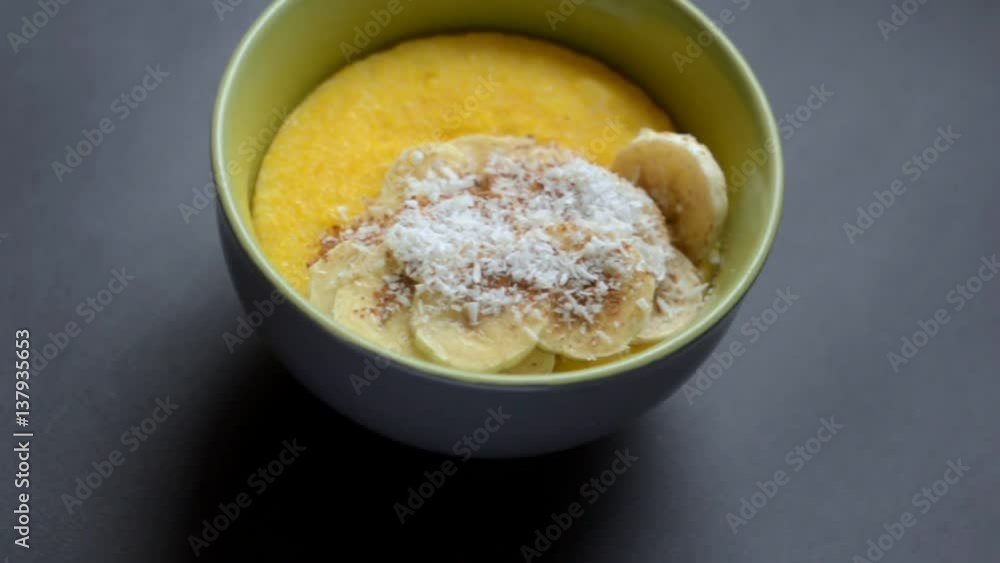 Corn porridge with banana, coconut and cinnamon in a deep green bowl on black background. Healthy breakfast concept. Slow slide right to left . Top view Corn porridge with banana, coconut and cinnamon in a deep green bowl on black background. Healthy breakfast concept. Slow slide right to left . Top view