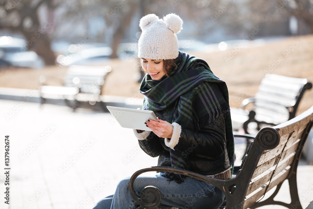 Smiling woman sitting on bench and using tablet outdoors