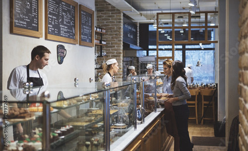 Two young women standing at bakery shop and chatting with baker.
