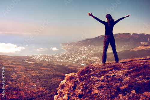 Cheering young woman standing at sunrise seaside mountain peak. Arms outstretched, freedom and happiness, achievement in mountains. Back view