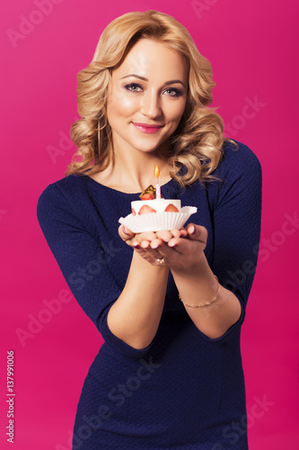Beautiful blonde woman in luxury blue dress and curly hairstyle blowing candle on birthday cake. clear skin. pink background