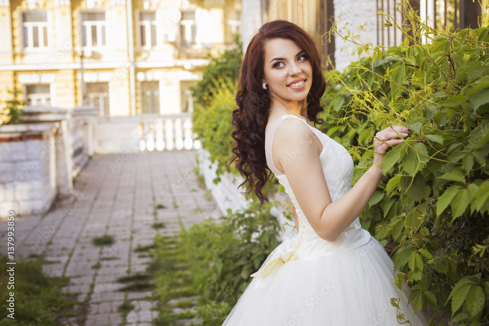 Beautiful brunette woman bride in a garden park in white wedding dress, curly hairstyle and a smile. warm weather, outdoors. copy space