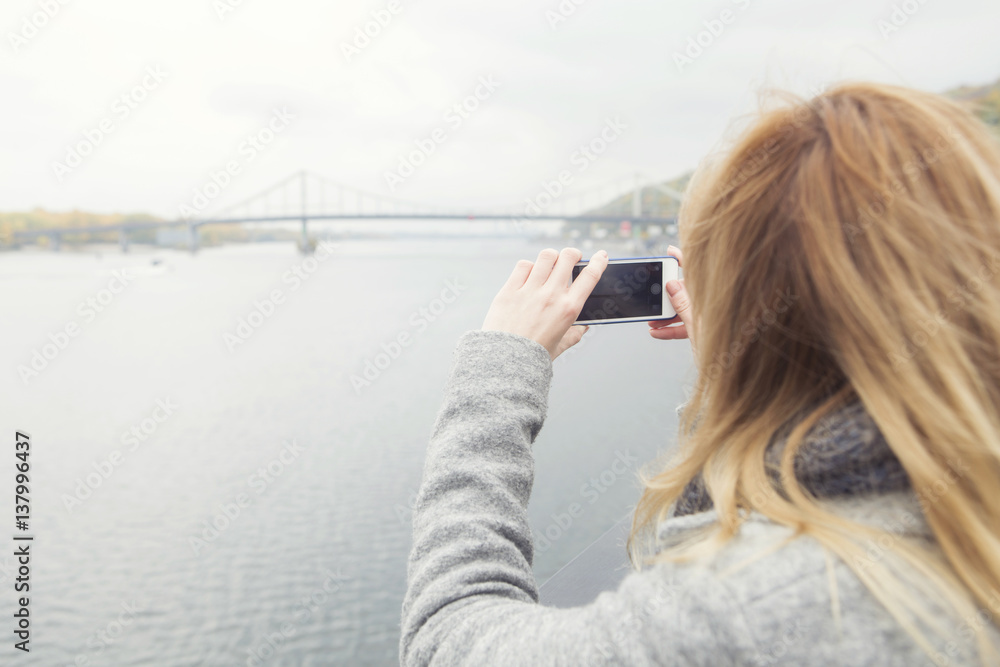 Blonde beautiful stylish caucasian woman in casual outfit on a walk in european city street  photographing view: bridge and river on her phone. copy space. focus on the phone