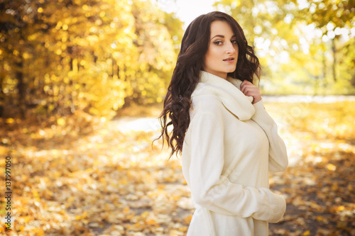 Beautiful caucasian brunette young woman in warm autumn day at park. Yellow leaves and sunshine. Fall season. Woman in white sweater, smiling. Copy space