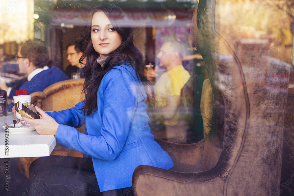 Brunette woman in business clothes: grey sweater and blue jacket sitting at the cafe near the window in european city drinking latte coffee and working using her tablet and smartphone. copy space