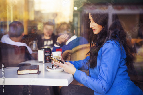 Brunette woman in business clothes: grey sweater and blue jacket sitting at the cafe near the window in european city drinking latte coffee and working using her tablet and smartphone. copy space