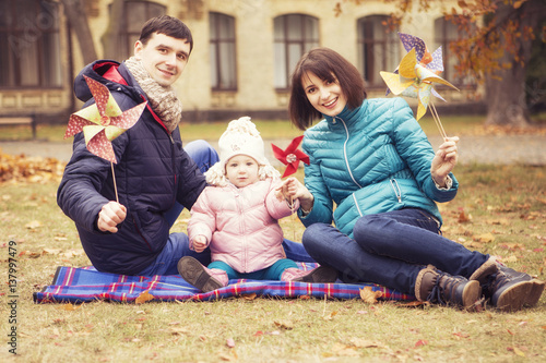 Happy loving family(mother, father and little daughter kid) outdoors walking having fun on a park in autumn season. Fallen yellow leaves on a background. Cold weather