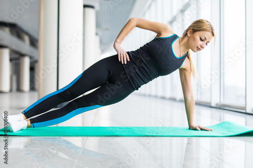 Young beautiful woman in sportswear doing side plank and looking at camera in front of window at gym