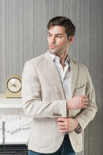 Man model in light beige linen jacket, white shirt and blue jeans near fireplace