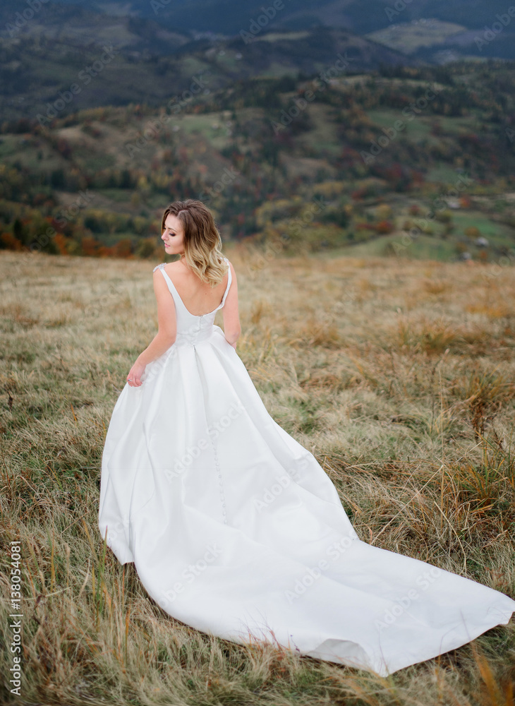 Bride in long dress with open back looks over her shoulder standing on the hill