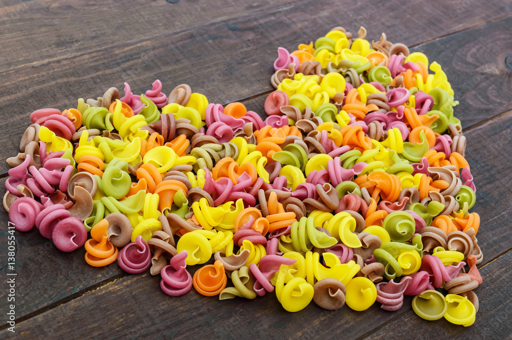 Colorful pasta colored by vegetables (beets, greens, spinach, carrots, tomatoes, peppers) on a dark wooden table in form of heart . Healthy food concept.