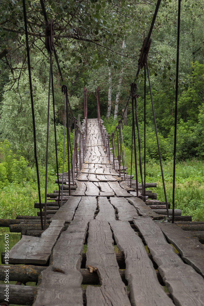 A suspended wooden bridge in green summer forest. Large cracks in the old boards and rusted cables of the suspension bridge. Forest landscape