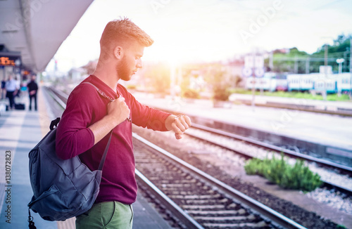 Young man checking the time on wristwatch at rail station platform - Student commuter waiting train at railway departures in pensive facial expression 