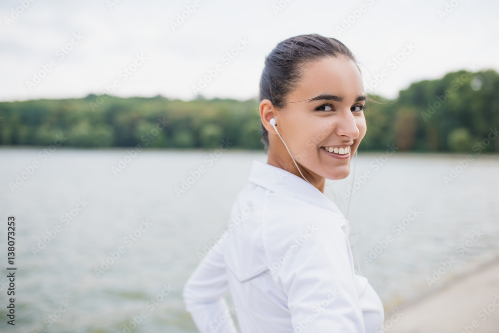 Portrait of young beautiful caucasian woman wearing sportswear looking at camera from back. Female athlete listening music with earphone. Smiling girl runner listen music in earphones from smartphone.