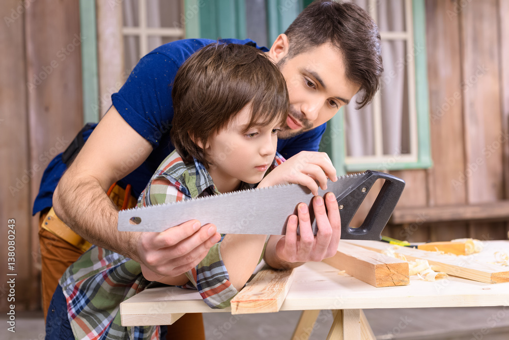 Pensive father and son leaning on wooden table and inspecting hand saw Pensive father and son leaning on wooden table and inspecting hand saw