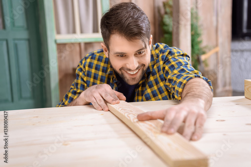 Smiling bearded carpenter measuring wooden plank on table