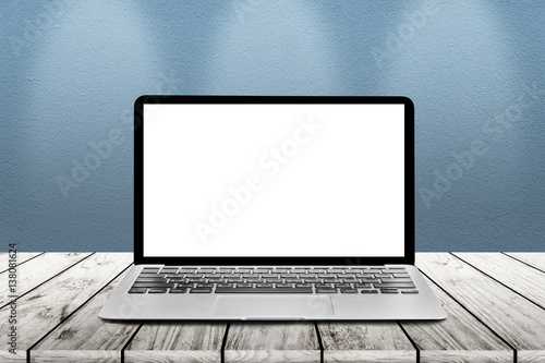 Laptop with white blank mockup screen is on top of wood table with concrete wall and light spot background.