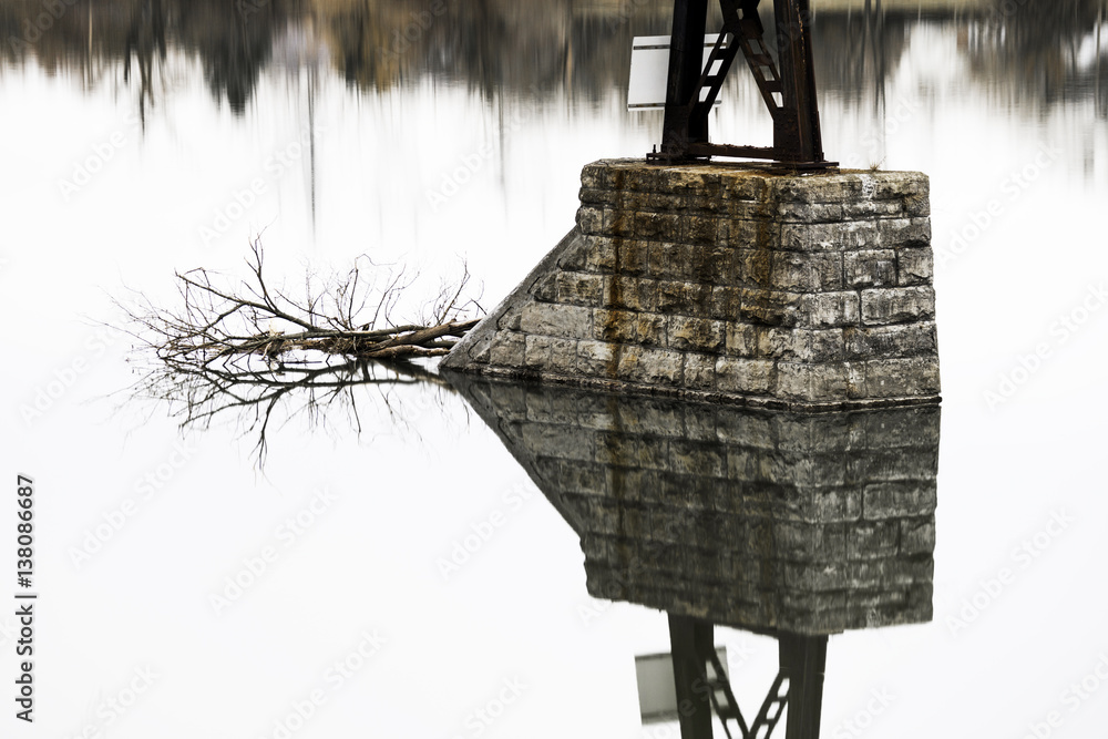 Mirrored reflection in perfect symmetry of bridge stone pillar and fallen branch on calm lake
