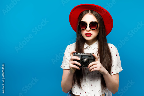 Fashion pretty young smiling woman holds retro camera wearing red hat, over blue background.