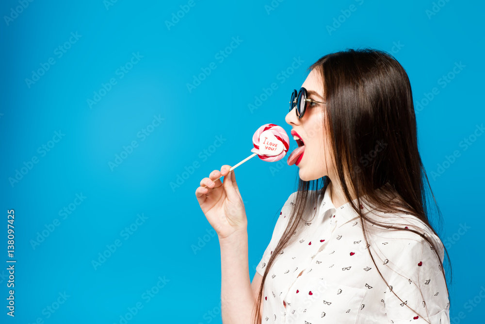 Young beautiful woman holding lollipop isolated on blue background. Happy girl wearing sunglasses eating multi colored candy. joyful and cheerful woman eating lollypop having fun.