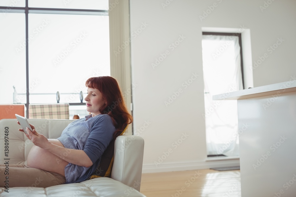 Pregnant woman digital tablet while relaxing on sofa in living room at home Pregnant woman digital tablet while relaxing on sofa in living room at home
