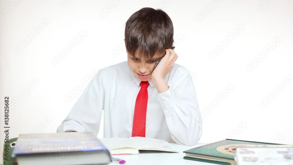 Young Caucasian school kid in red tie sitting at table writing talking on mobile phone on white background in slowmotion Young Caucasian school kid in red tie sitting at table writing talking on mobile phone on white background in slowmotion