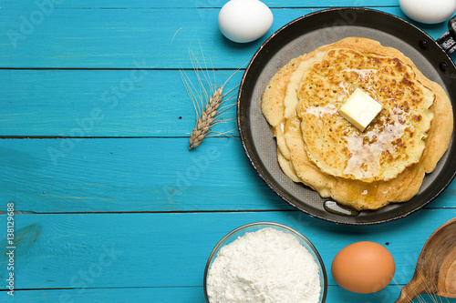 Hot delicious pancakes in frying pan on blue wooden table with flour and eggs