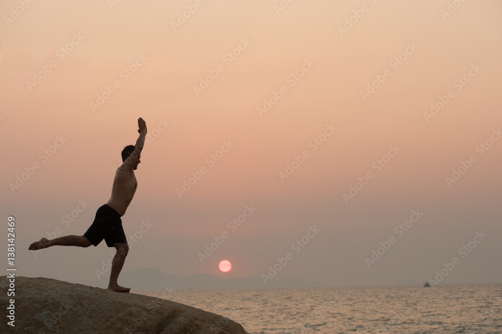 Male is engaged in fitness yoga exercise on the stone in Sunset.