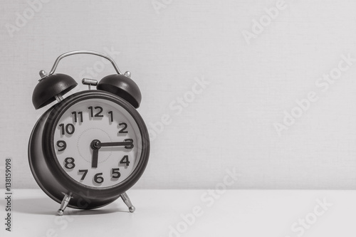 Closeup alarm clock for decorate show a quarter past six or 6:15 a.m.on white wood desk and cream wallpaper textured background in black and white tone with copy space