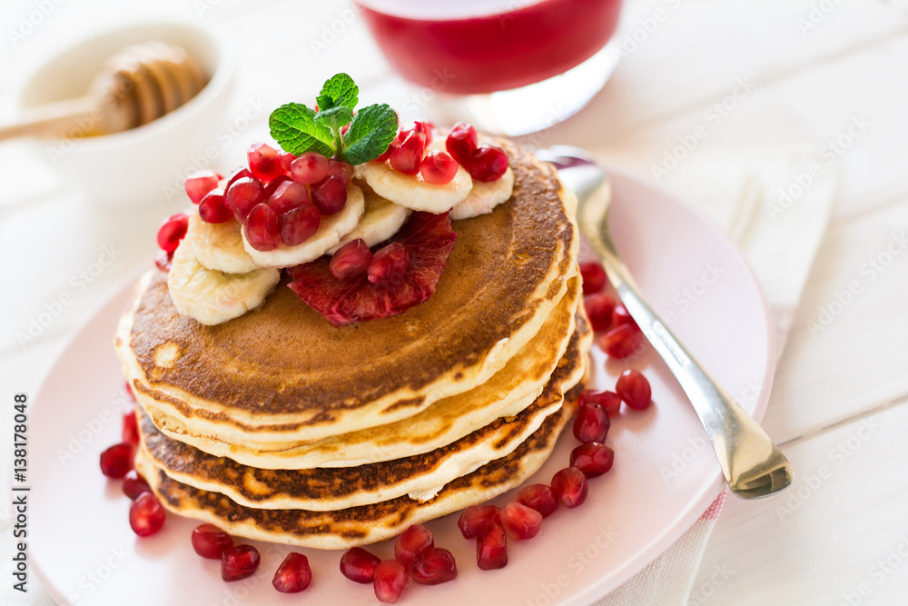 Traditional breakfast: stack of pancakes with banana slices and garnet seeds on white wooden table. Selective focus Traditional breakfast: stack of pancakes with banana slices and garnet seeds on white wooden table. Selective focus