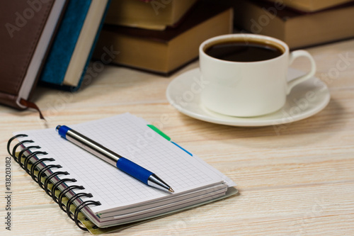 Notebook with pen on a wooden table in front of the window. A cup of hot coffee on the table. A stack of books.