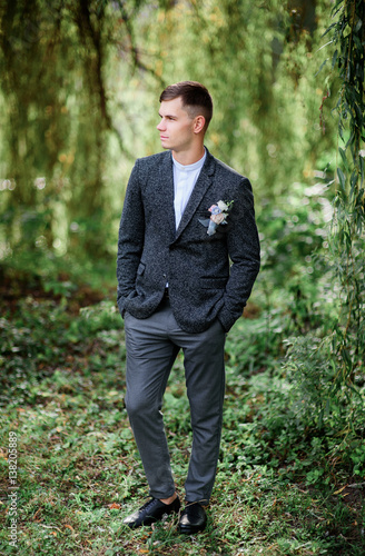 Thoughtful groom poses in grey suit before green bushes