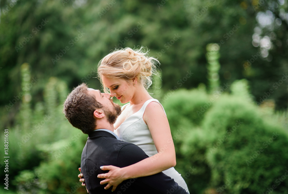 Bearded groom raises blonde bride up posing on green lawn in park Bearded groom raises blonde bride up posing on green lawn in park