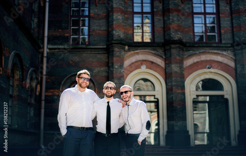 Groom and groomsmen in black sunglasses hold their hands on each other shoulders posing on the backyard of an old house