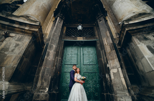 Tall groom hugs bride from behind standing with her before entrance to an old cathedral © IVASHstudio